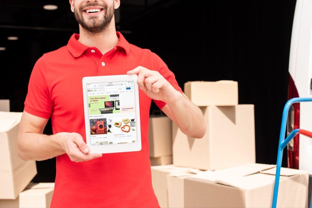 Smiling mover in red shirt holding a tablet in front of moving boxes, representing how moving companies can improve visibility with local SEO services.