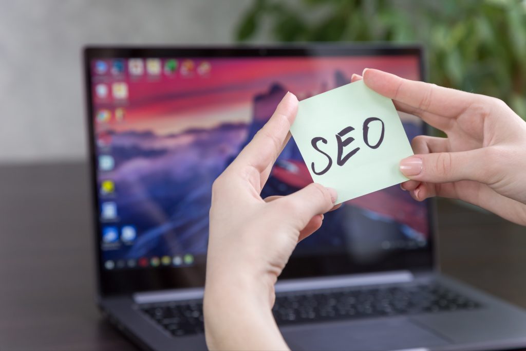 Hands holding a sticky note with "SEO" written on it in front of a laptop screen.